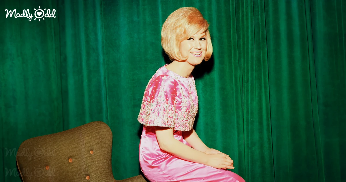 Dusty Springfield sitting on chaise with green curtain background behind her.