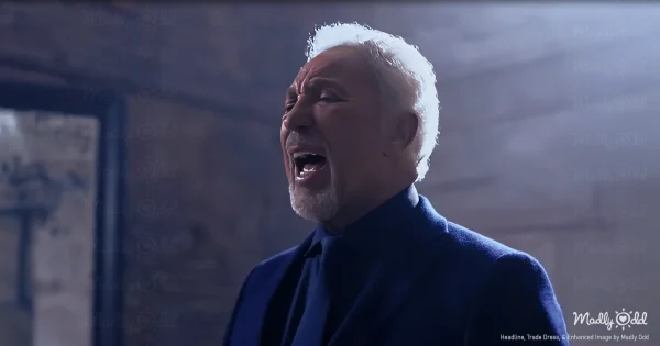 A dimly lit room with Tom Jones sitting alone in a chair, old black-and-white film clips of Elvis playing on a wall in front of him. The camera captures his weathered face as he sings, the strain of emotion evident in his expression.